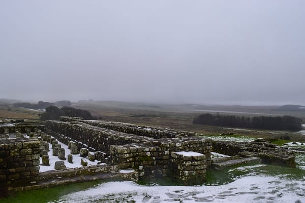 The remains of Housesteads Roman Fort with partial stone structures still standing, with a light dusting of snow. Visit the fort on day 1 of this Northumberland itinerary.
